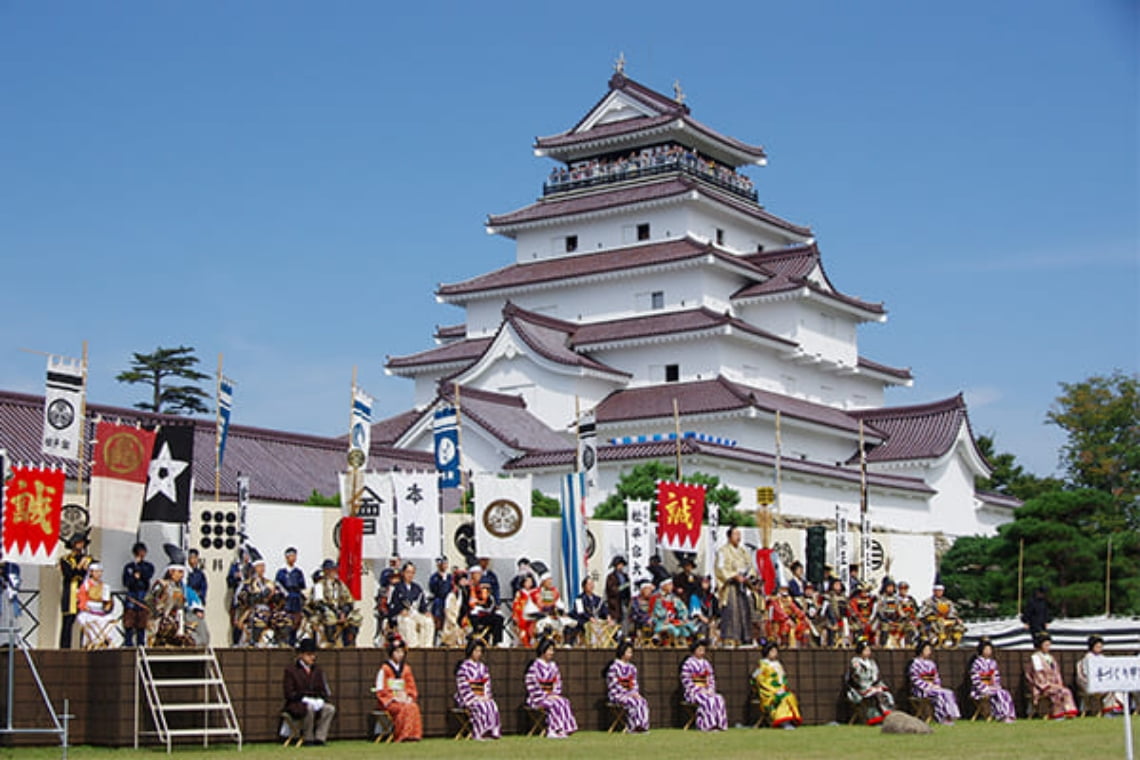 地域のお祭りで、城を背景に伝統衣装をまとった参加者が行列や演舞を行う様子